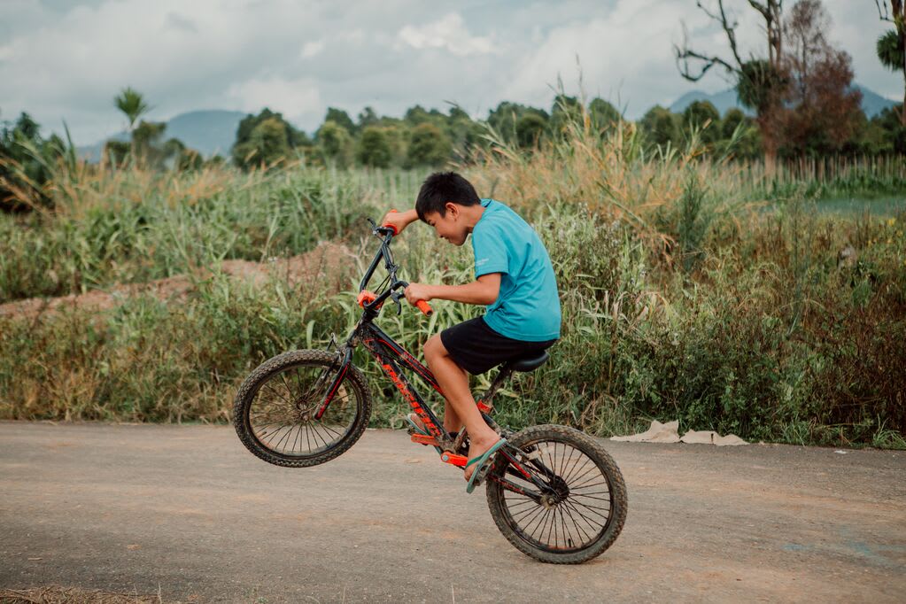 Little boy wearing a blue t shirt pulls up the handlebars of his bike so the wheel is in the air.